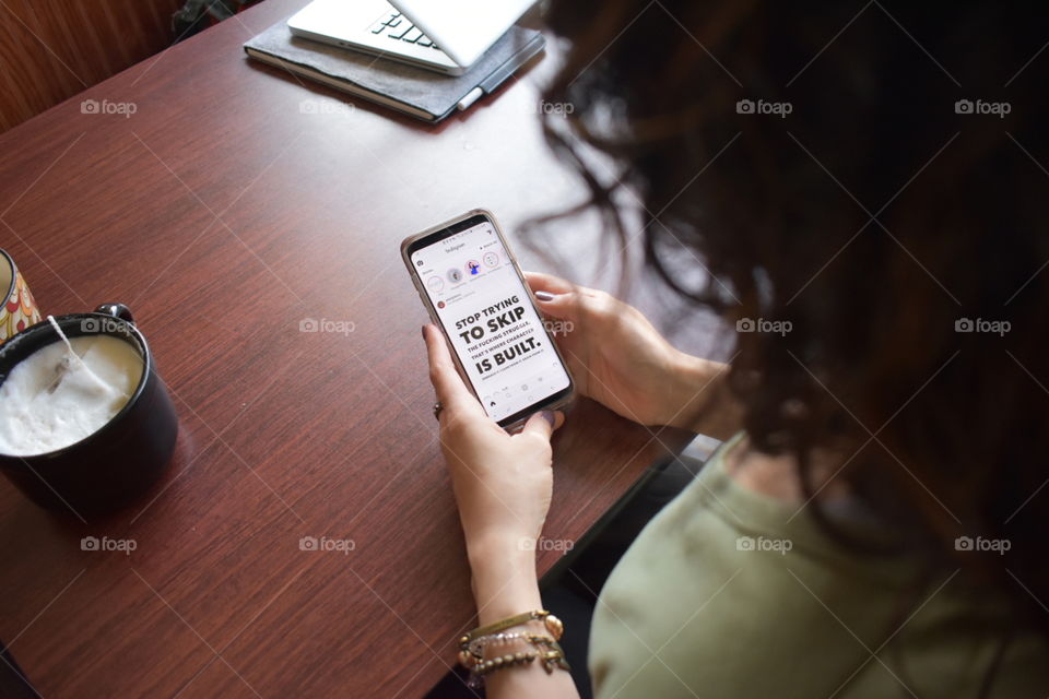 woman sitting at a table using her cell phone to browse Instagram. bracelets. computer.