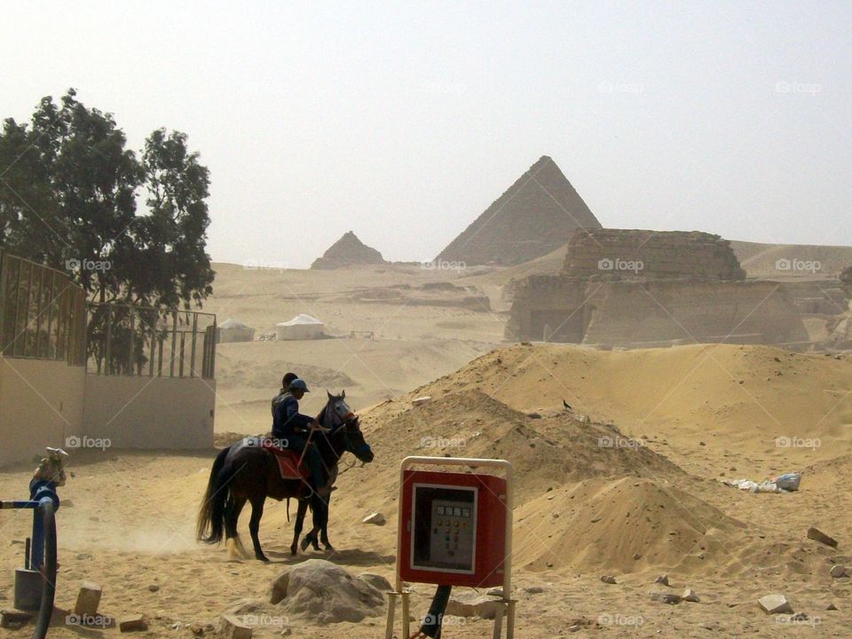 Two horseback riders near a water station in the foreground of the ancient pyramids 