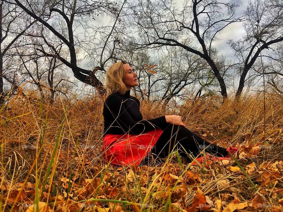 девушка в красной юбке сидит на жнлтой листве на фоне деревьев и неба
