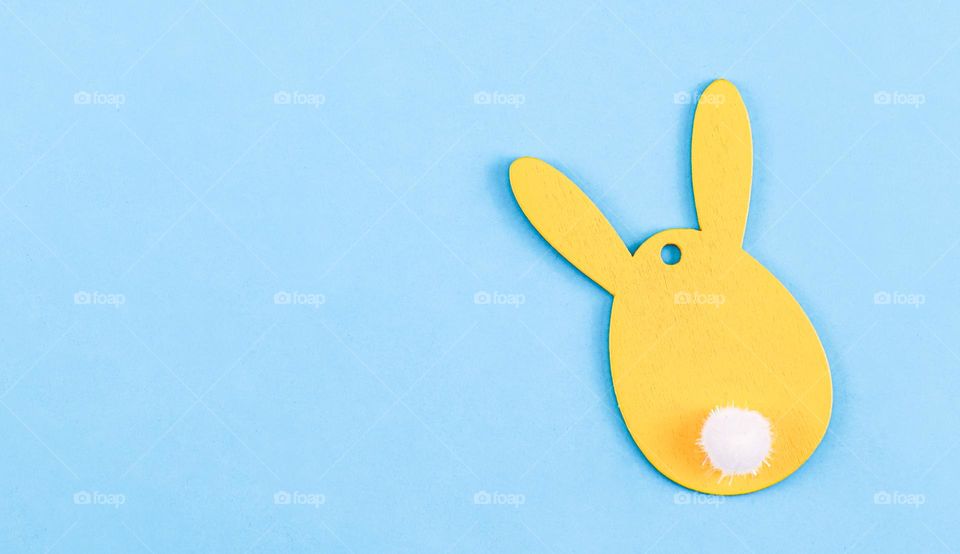 One creative yellow wooden easter bunny with a fluffy white tail lies side by side on a blue background with copy space on the left, flat lay close-up. Easter decoration concept.
