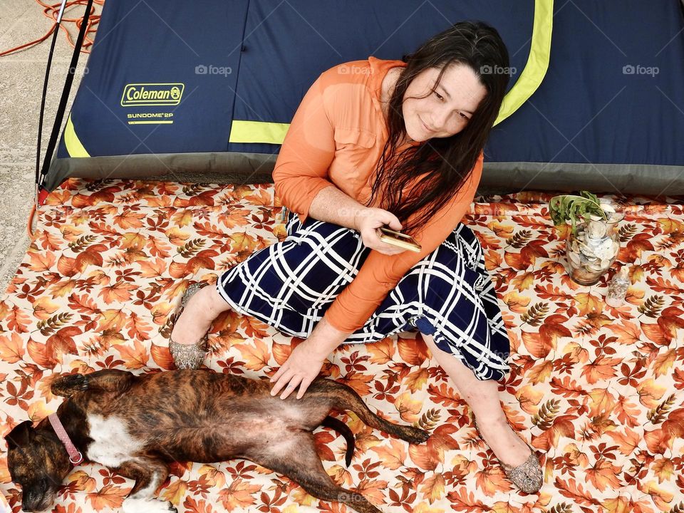 Autumn fall decor season during camping, woman sitting on a step on a cloth with orange leaves with blue and orange clothes in front of a blue and green tent and holding a phone and petting her cute brindle dog Bella laying on her back.