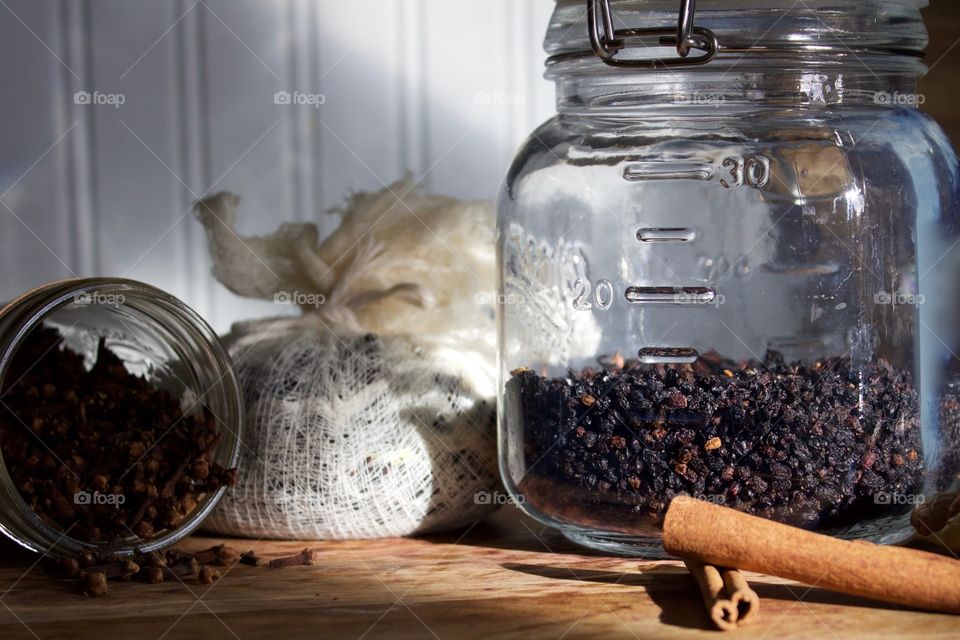 Ingredients for elderberry syrup—cloves in small glass jar, elderberries and chopped ginger in cheesecloth, dried elderberries in sealed glass jar and cinnamon sticks on wooden surface