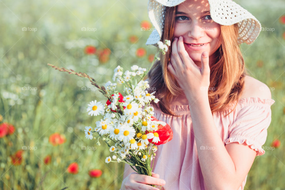 Beautieful young girl in the field of wild flowers. Teenage girl picking the spring flowers in the meadow, holding bouquet of flowers. She wearing hat and summer clothes. Spending time close to nature