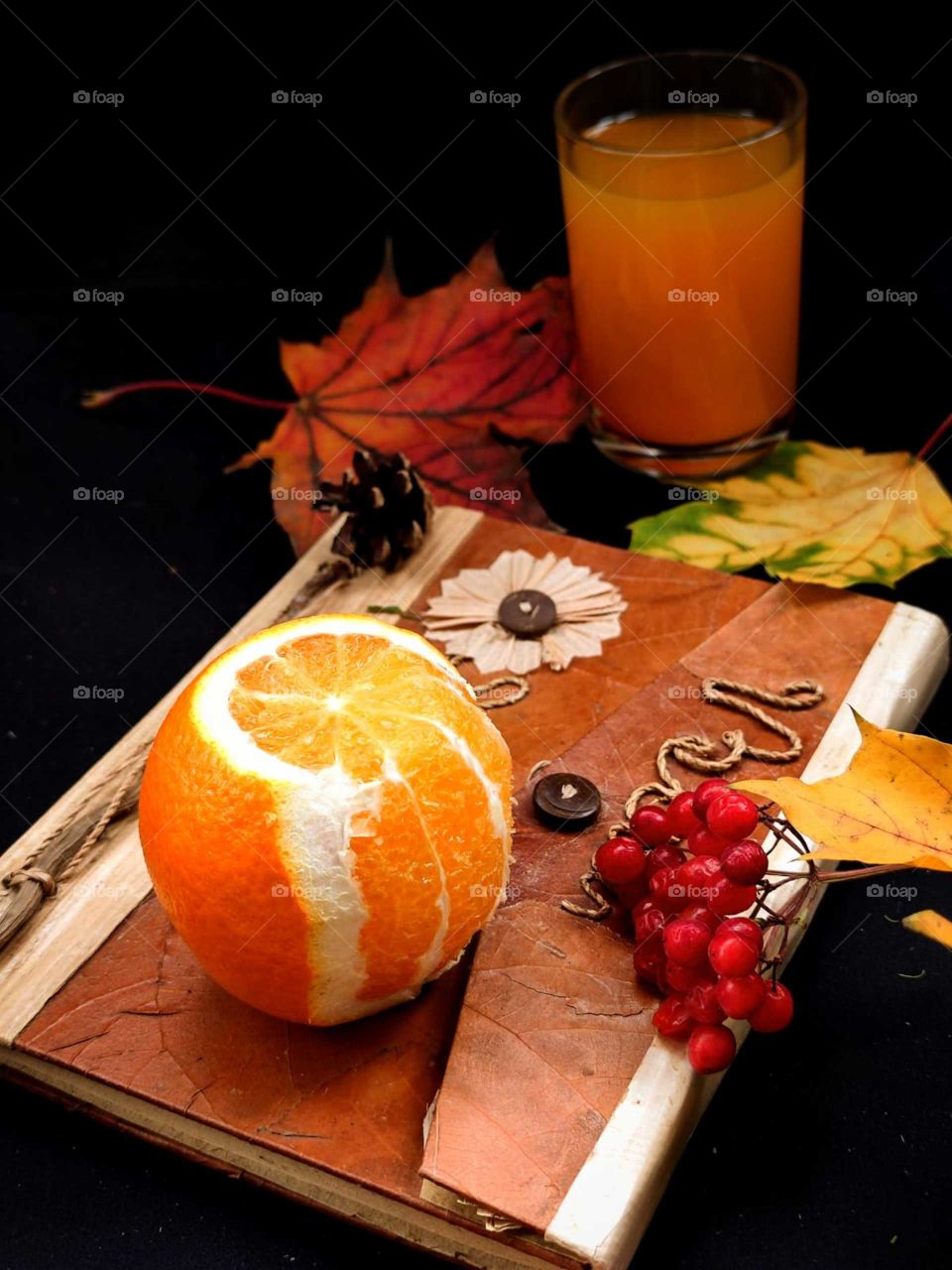 Autumn composition.  Book.  Half peeled orange and red rowan berries.  Next to the book are a red maple leaf, yellow maple leaves.  In the background is a glass of orange orange juice.  Black background