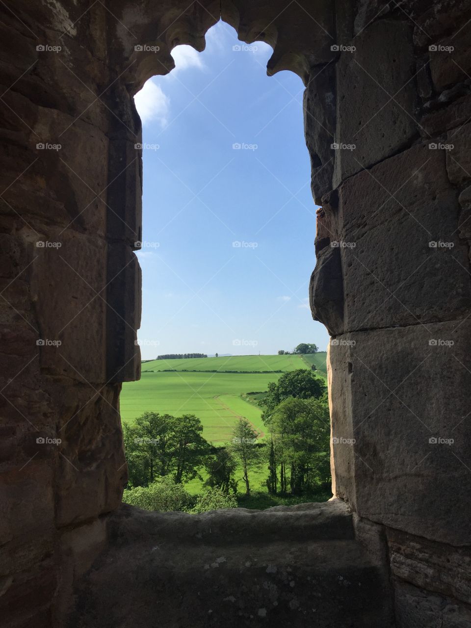 Looking through the remains of an old window at Raglan Castle, Wales. In the distance, luscious green grass with clear blue skies.
