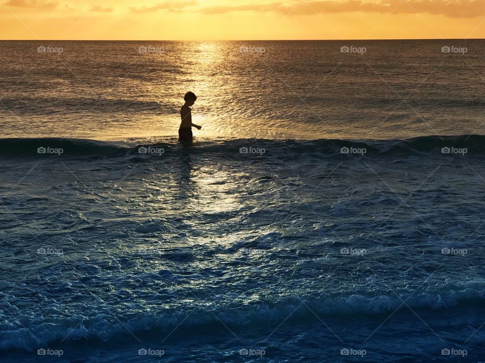 Small boy walking through the ocean as a golden sunset bathes him in light.