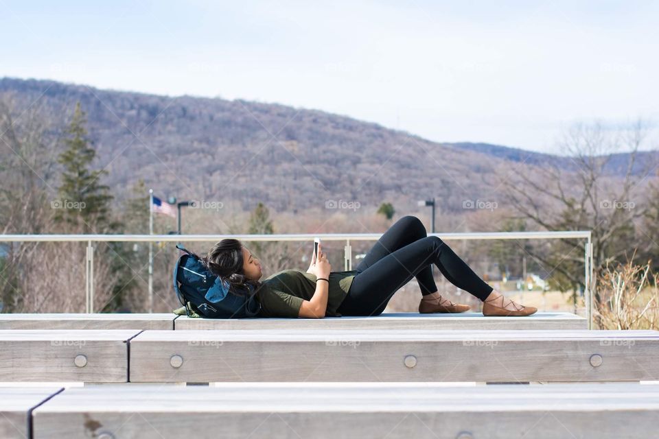 A woman uses her phone while relaxing on a bench by mountains 