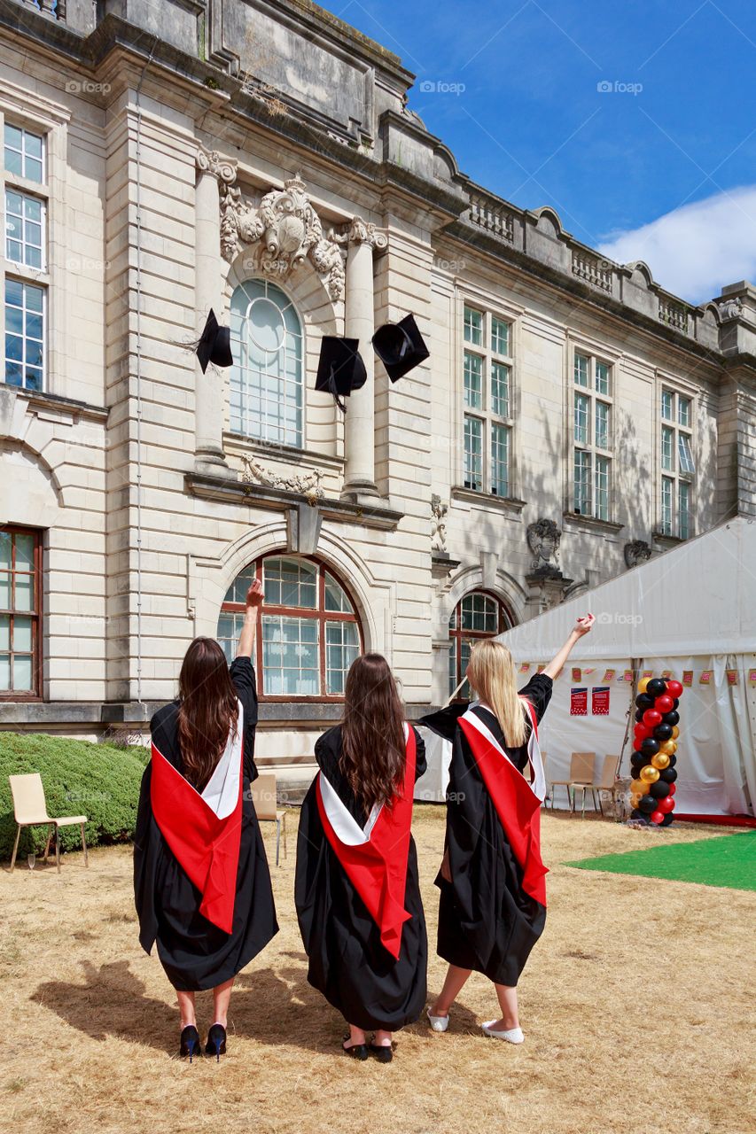 Back side view of three happy girls wearing dresses throwing academic caps in yard of Cardiff University