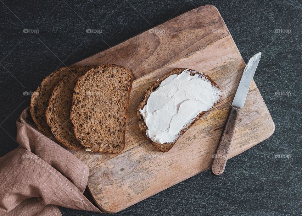 Torn pieces of bread with sunflower seeds spread with cheese mass on a wooden cutting board with a kitchen napkin and a knife lie diagonally on a chain stone background, flat lay close-up. The concept of baking bread, a healthy breakfast.