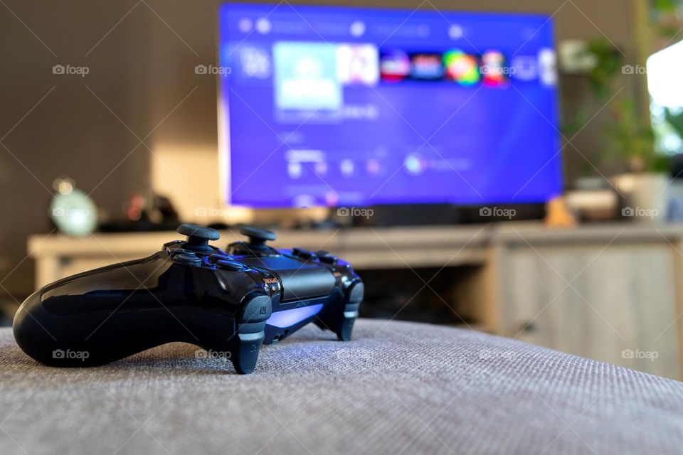 a portrait of a Playstation 4 wireless controller lying on a couch in front of a tv showing the playstation home screen.