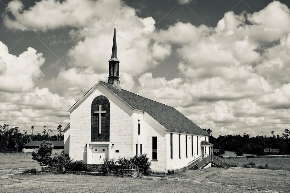 A black and white picture of a white church with a steeple, a cross, windows, and steps with puffy white clouds in the sky