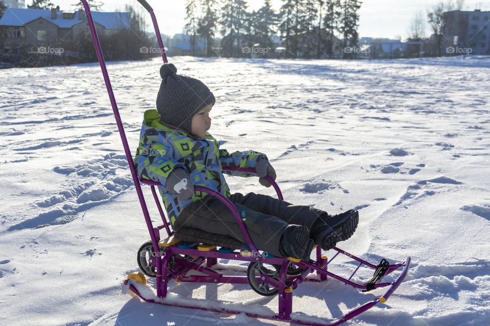 A child with a serious expression on his face in winter clothes jackets, pants, hat and boots in winter on white snow on the street and in the park in nature sledding and playing winter fun.