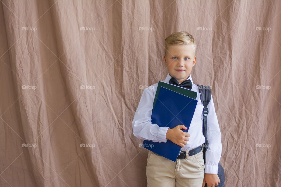 Smiling blond schoolboy in white shirt with bow tie backpack and notebooks return to school copy space  