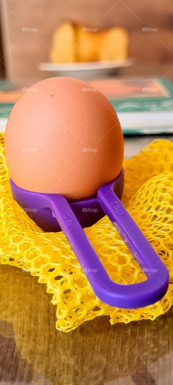 Purple kitchen utensil with an egg inside, wrapped in a yellow ecological mesh bag.  In the background, a blurred image of a cookbook and a homemade cake.