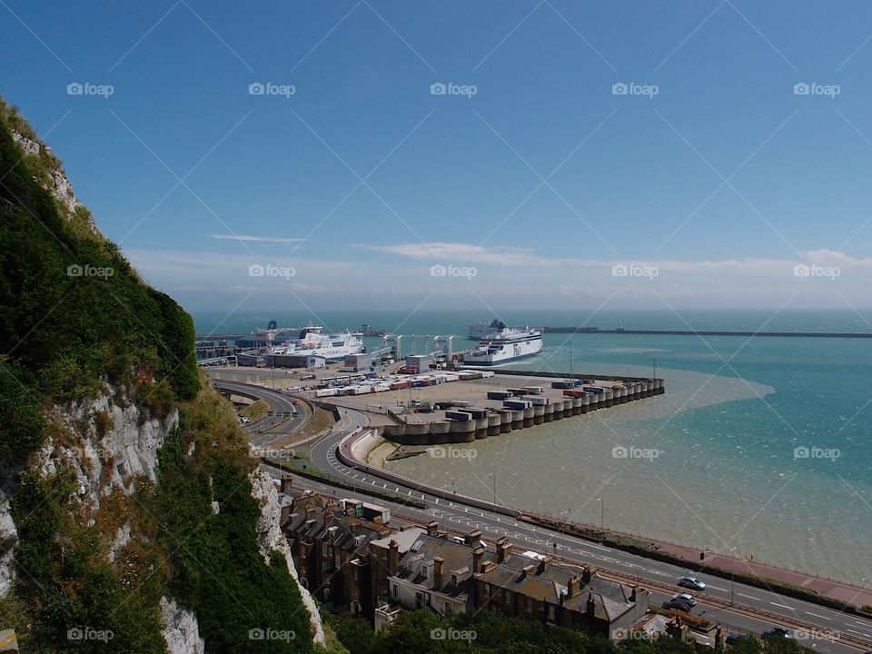 A road winds around a hill next to an ocean bay with a large ship on a sunny summer day in Folkestone, England. 