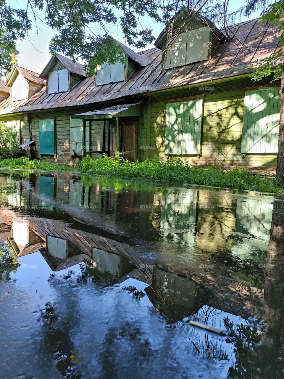 An old wooden house with boarded up windows stands among the trees.  An old house, green trees and a blue sky are reflected on the wet pavement.