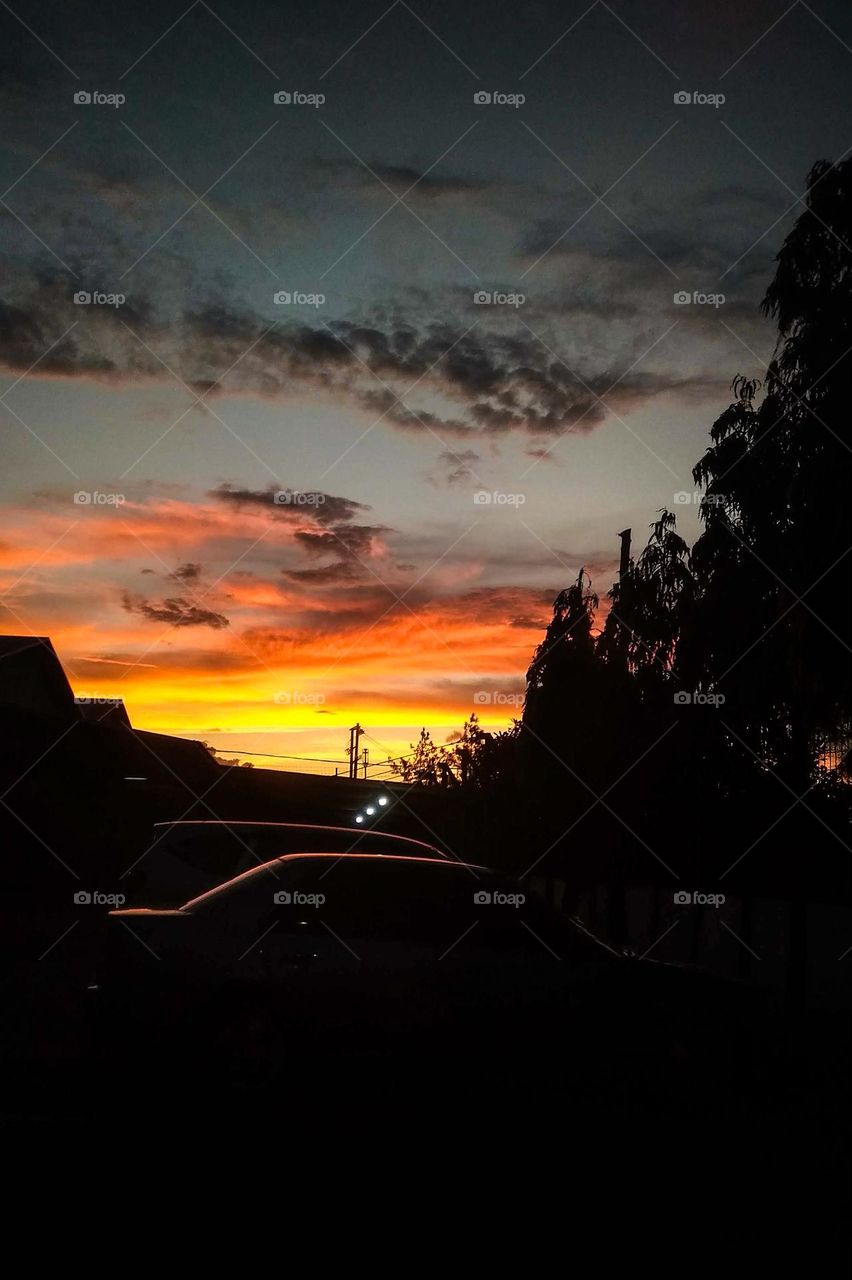 a beautiful sunset scene with a dark orange and blue sky, tree silhouettes, and a car