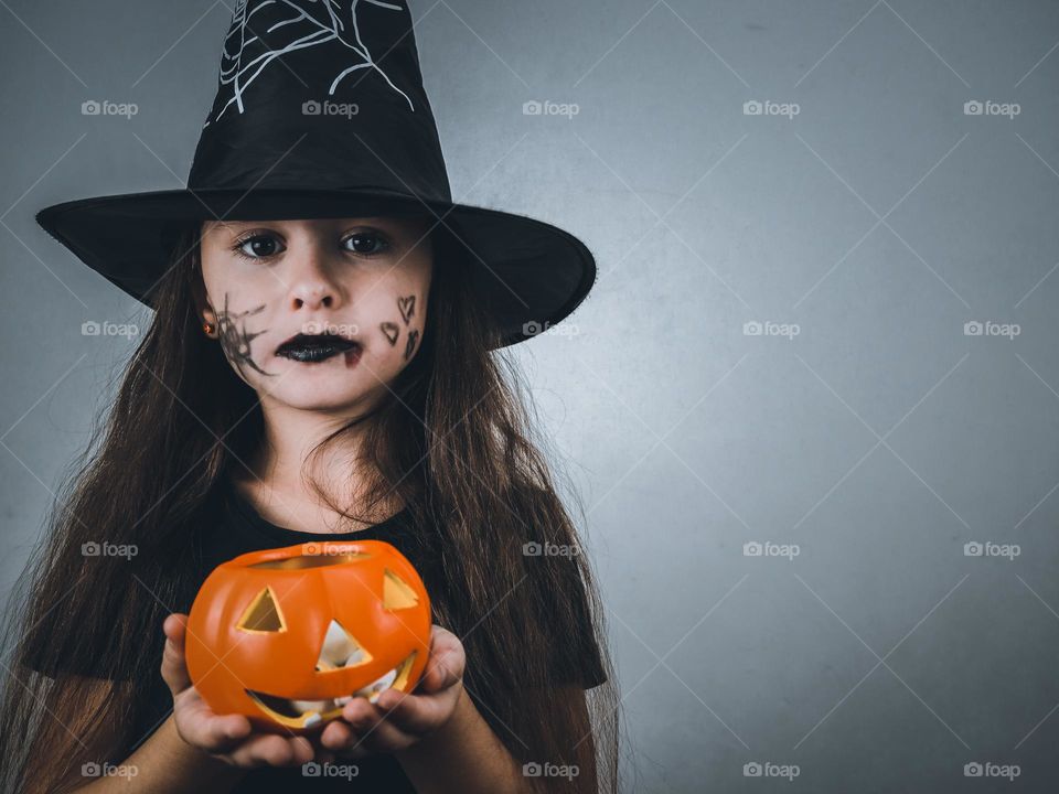 Portrait of a beautiful caucasian girl in a witch costume with a hat and make-up on her face holding a decorative pumpkin with candies looking at the camera on the left on a dark gray background with copy space on the right,close-up side view.Hallowe