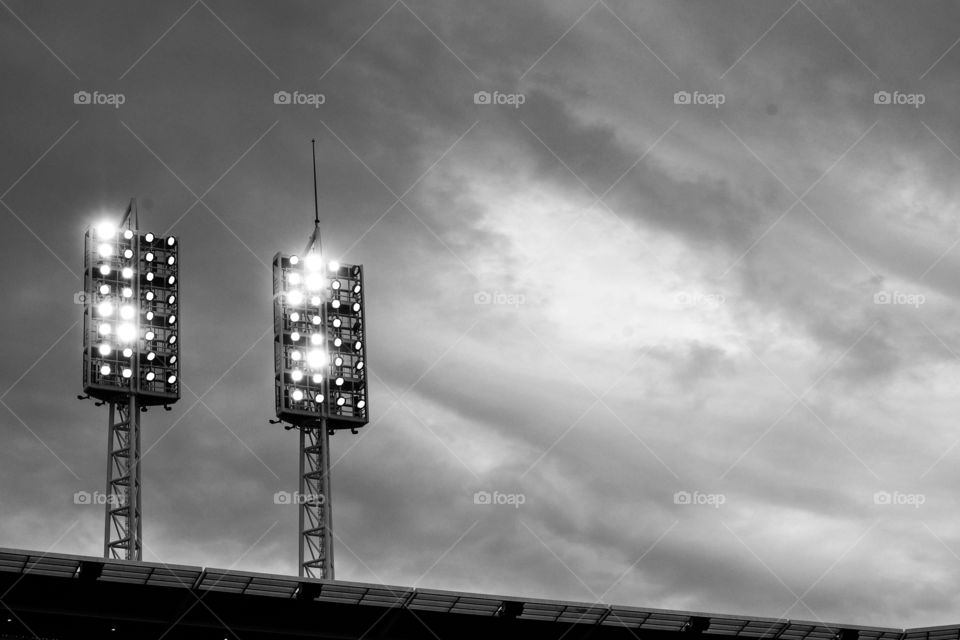 Black and white minimalist image of lit stadium lights against a cloudy sky