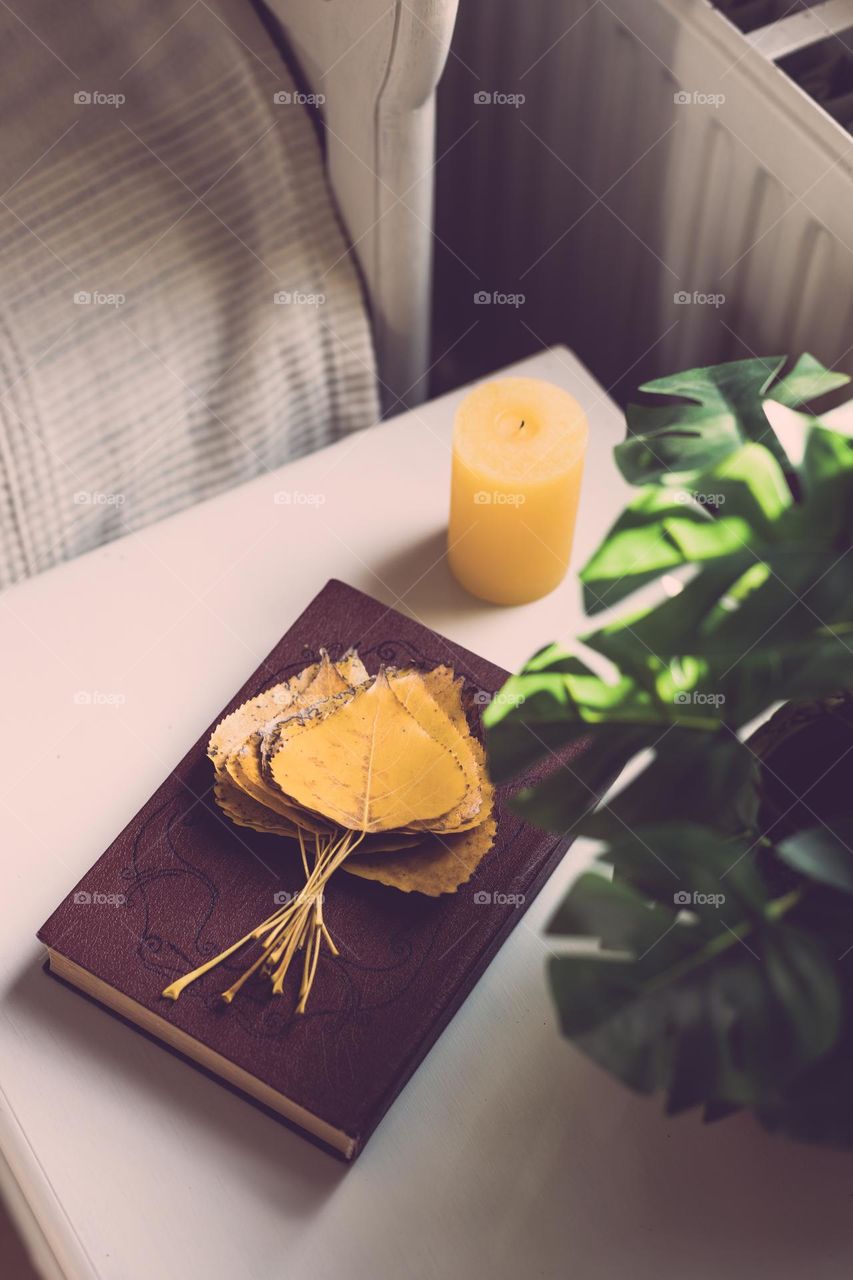 A bouquet of yellow poplar leaves on a brown book and a candle stand on the bedside table next to the bed in the bedroom in the early autumn morning, close-up side view.