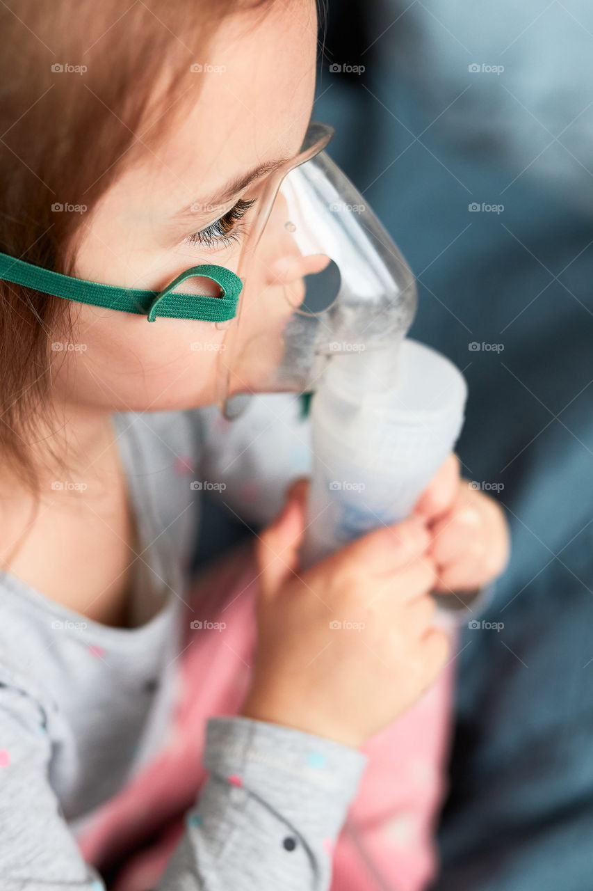 Sick little girl having medical inhalation treatment with nebuliser. Child with breathing mask on her face sitting in bed