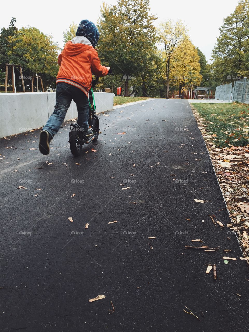 Boy on scooter in autumn park. Child from behind, riding scooter on track in park in rainy fall weather