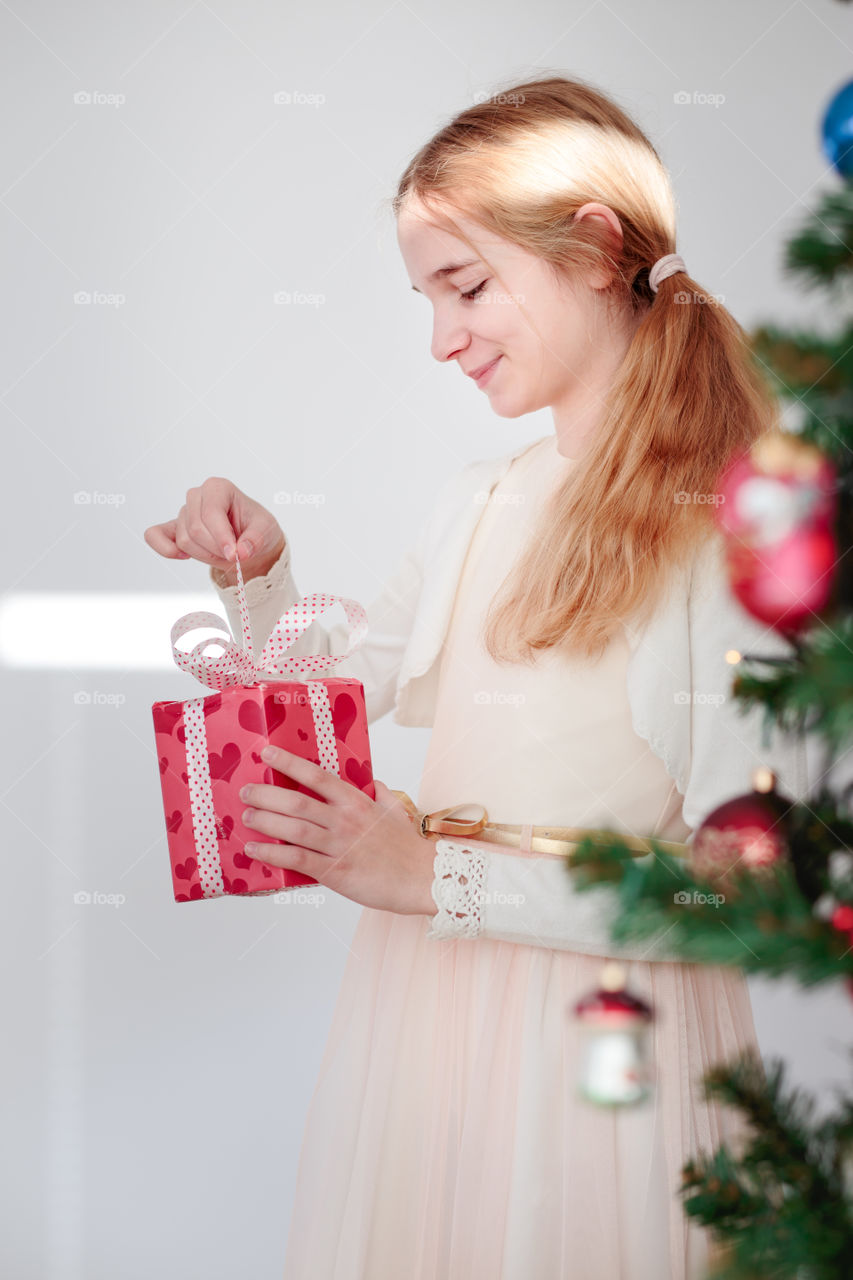 Happy smiling girl wearing pink dress standing behind a tree and  unpacking Christmas gift