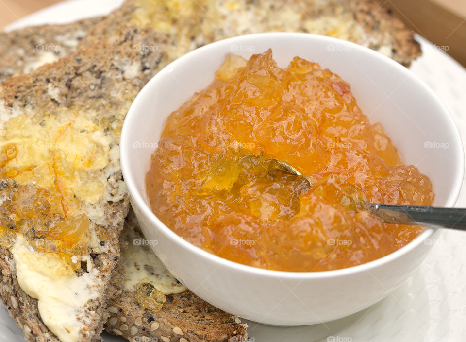 A white bowl filled with marmalade and a spoon beside buttered toast spread with marmalade.