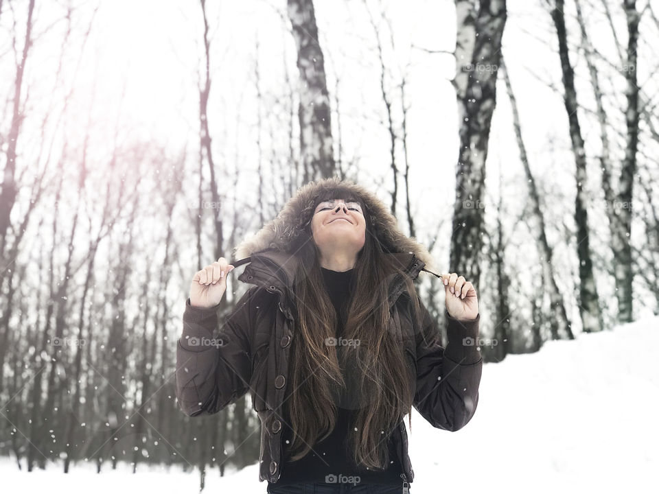 Young woman happily smiling under the falling snow in the forest 