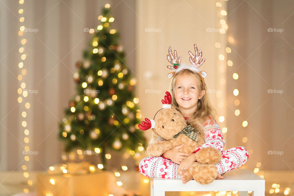 Cute little girl with blonde hair in pajamas sitting near Christmas tree with teddy bear 