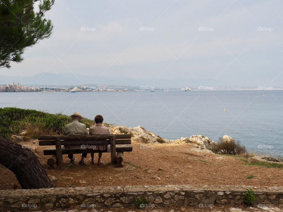 Cute elderly couple sitting on a bench looking out at the view of the coast of Antibes in the south of France.