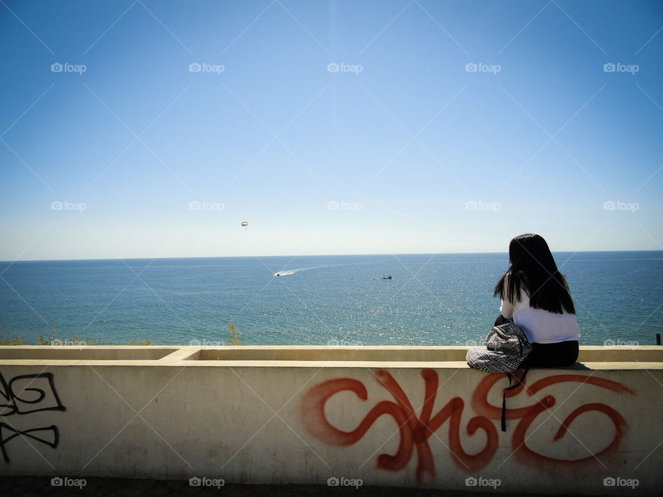 A girl sitting on a wall, looking over the ocean like she is waiting for the one she loves.