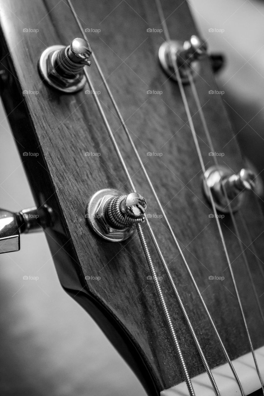 A black and white close up of the head of a wooden acoustic guitar, showing silver knobs and strings.