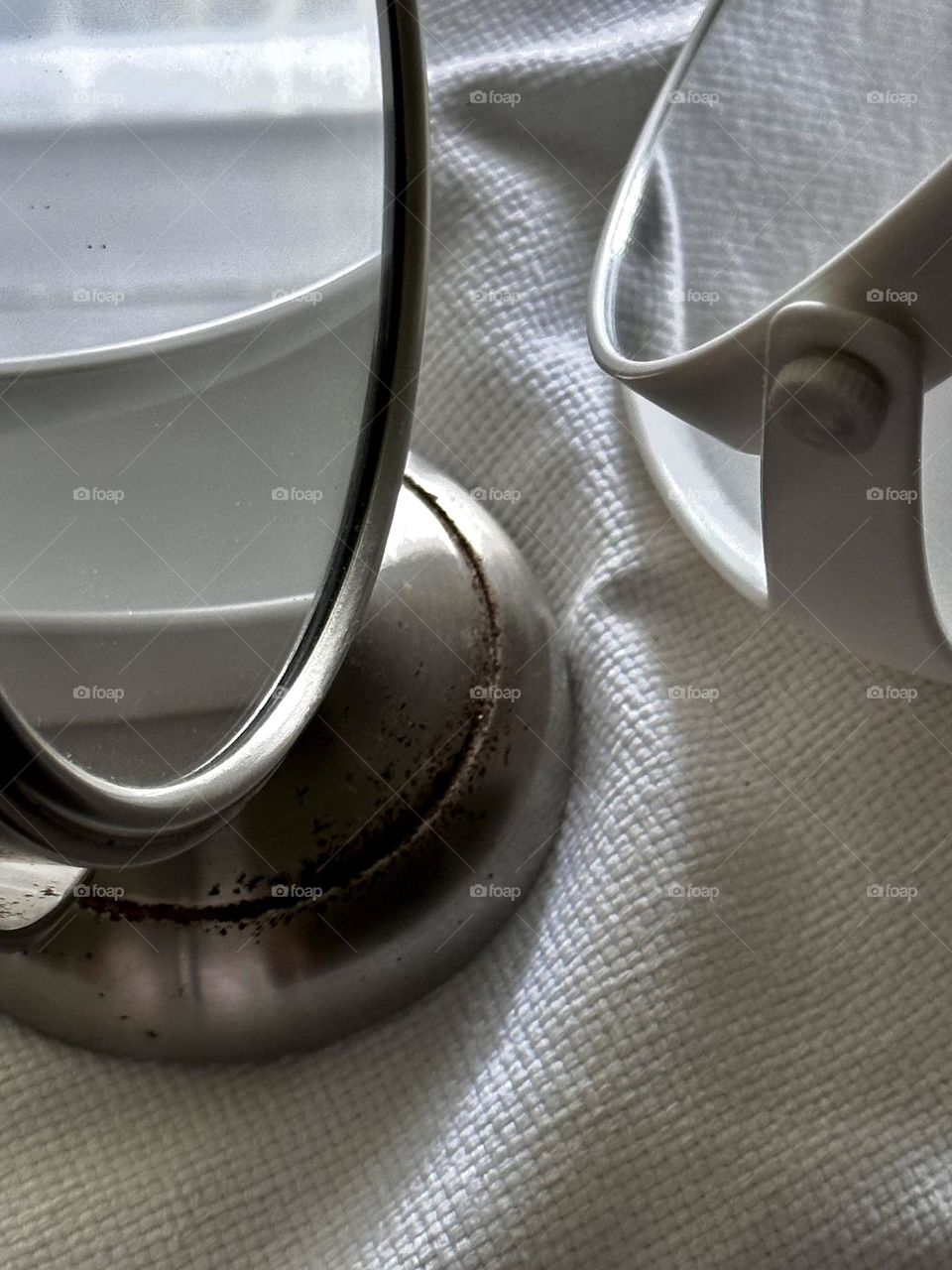 Two table top mirrors one white plastic and the other vintage stainless steel reflecting each other and the light on a white woven cotton cloth