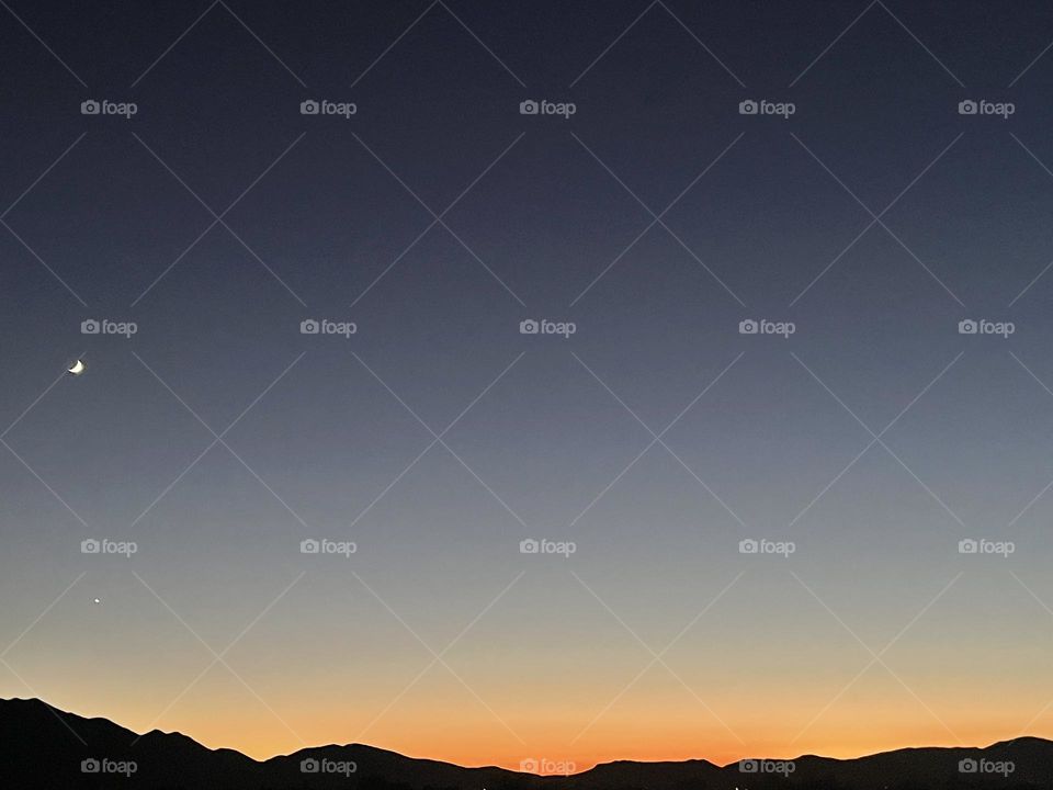 A orange and yellow sunset with mountains and the moon. 