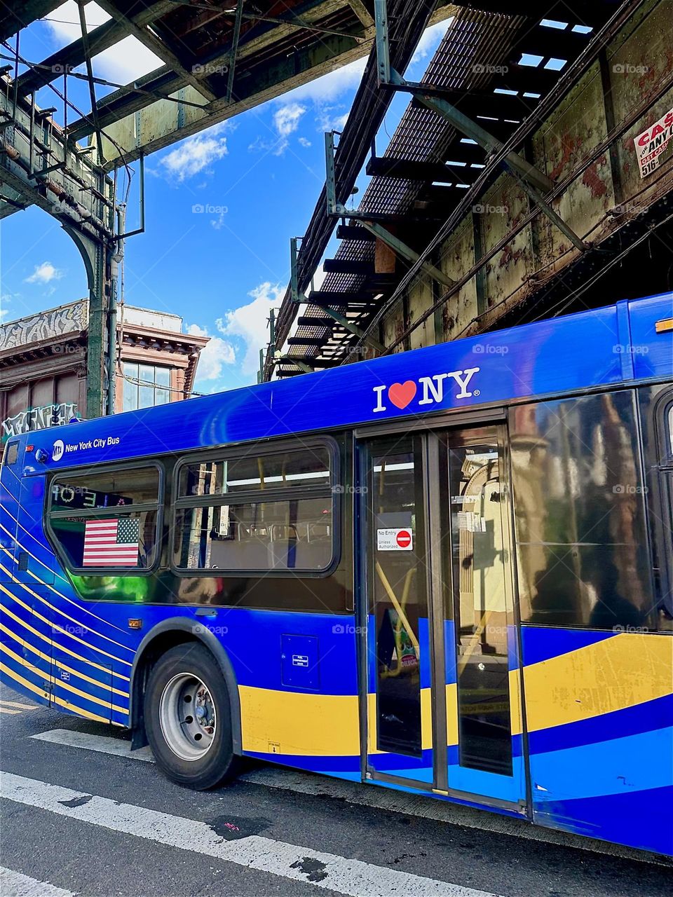 “I love NY” is written in white, the word “love” expressed by a red heart on the indigo background of a NYC bus that passes beneath two diagonal overpasses, one on “Myrtle Ave”, the other on “Broadway” in “Bushwick”, Bklyn. 2024. Hypnotic Productions