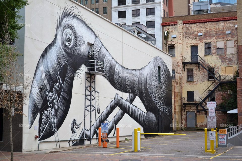 A black and white mural on a city wall of a bird-like creature and working men on stairs next to a brick wall