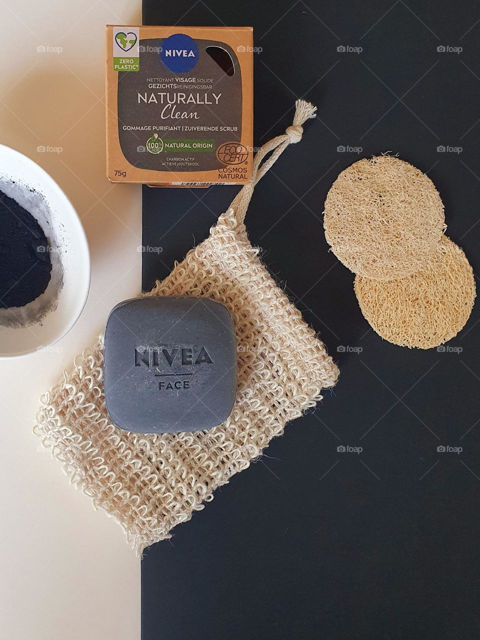 Flatlay on ivory and black papers of Nivea Naturally Clean purifying scrubbing soap placed on natural fibre pouch and box, natural fibre scrubbing pads and bone china cup containing charcoal powder