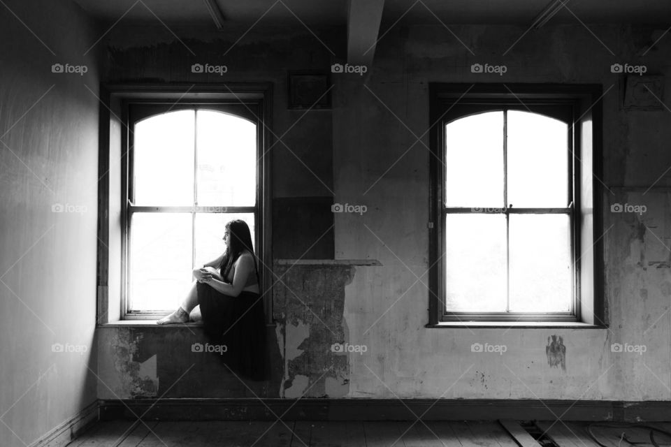 Girl sitting on the window bottom in a derelict building. She is silhouetted against the window. She is lit by natural light