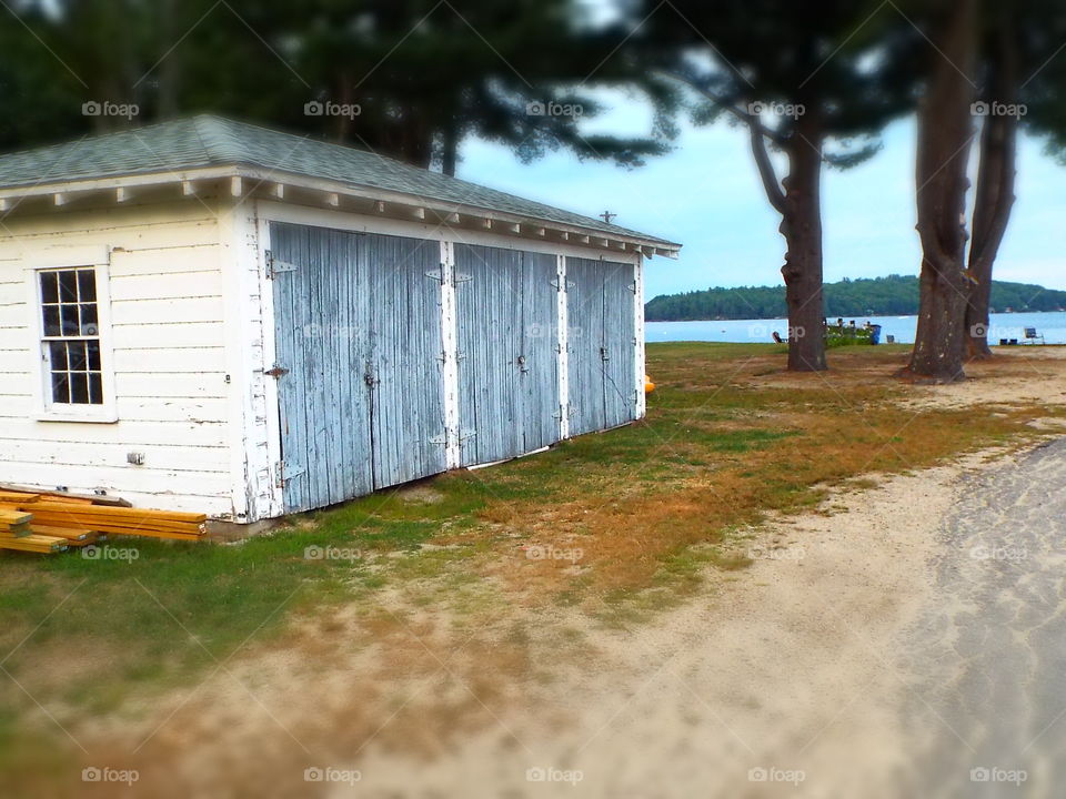 Old weathered wood equipment shed with peeling paint and lake and tall pine trees in background 
