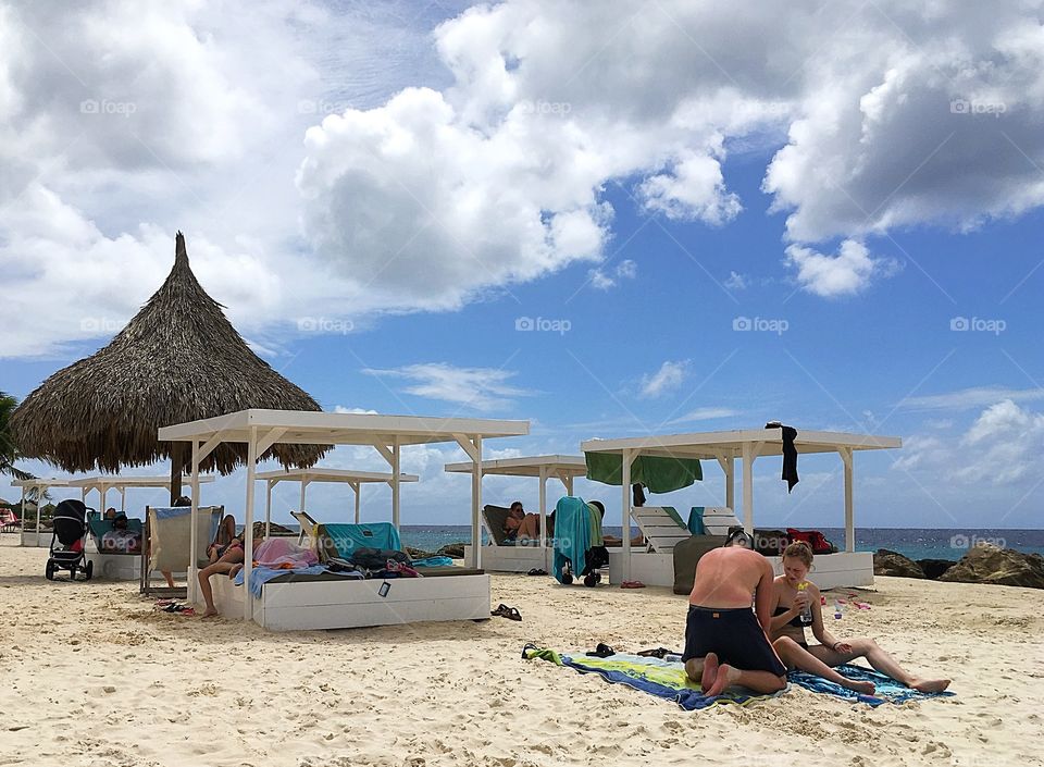 Candid photo of real people at the beach.  Daybeds and palapas on the beach with a backdrop of blue sky and white puffy clouds