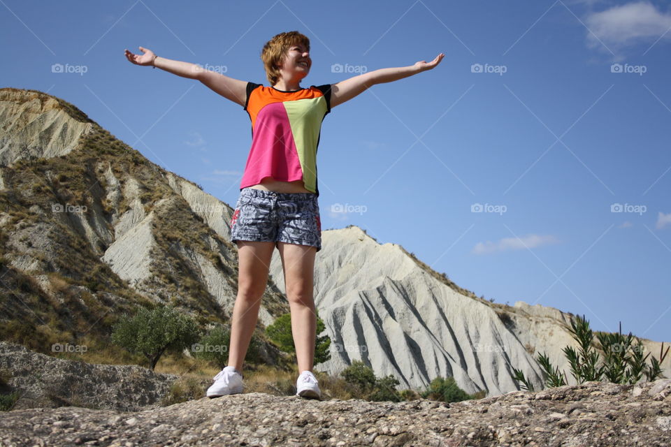 The main object is the model. Behind the model there is a beautiful mountain. The model is on a big rock. Around there are plants and trees.