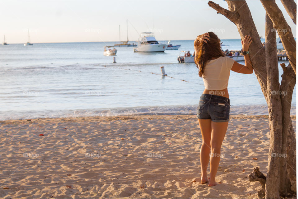 Girl leaning against tree facing the beach with boats 