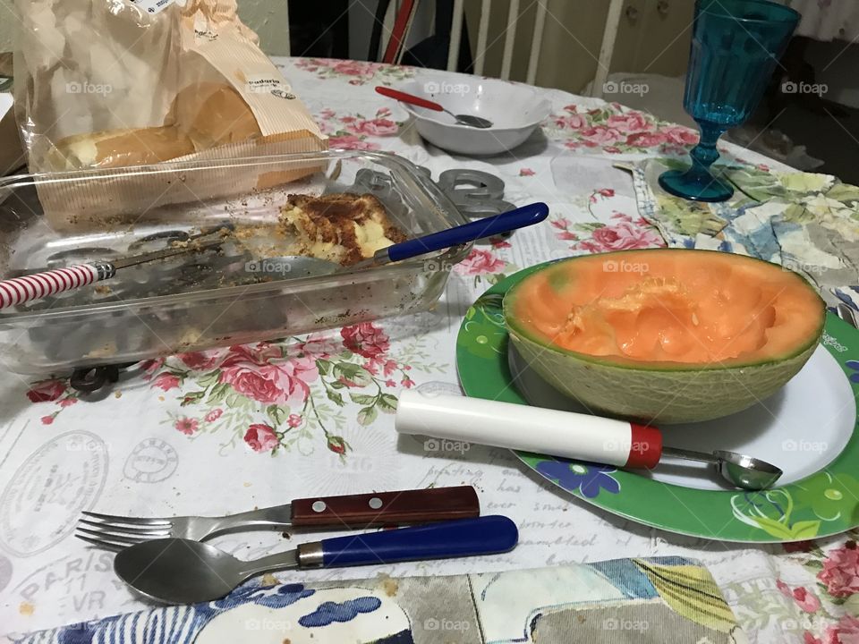 A table with spoon, fork, there’s an open mellon and a device to make balls. Also there’s a pice of pie and some bread over the table 