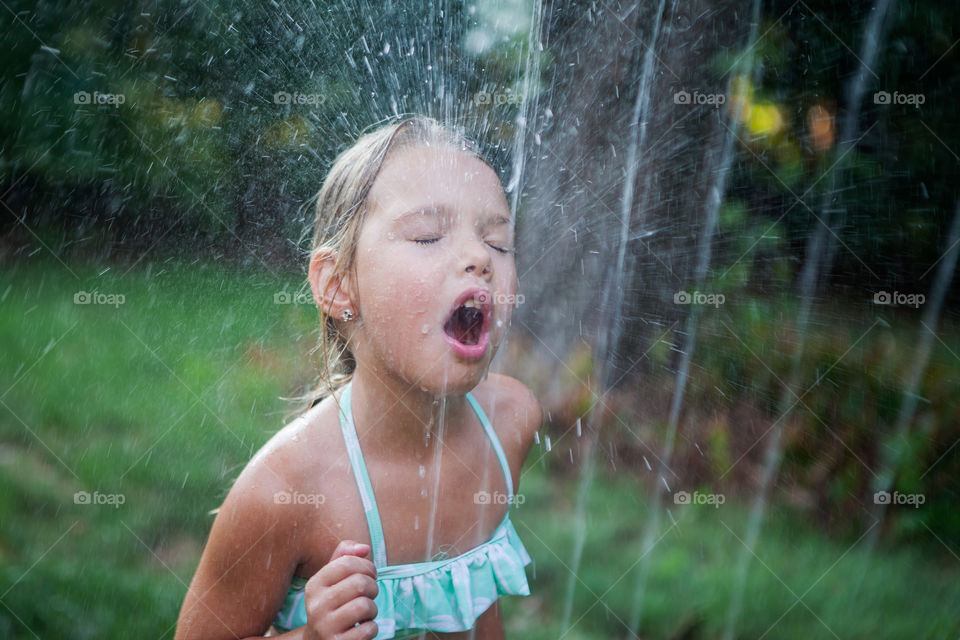 Cute little blonde girl drinking water from the backyard sprinkler during summer. 