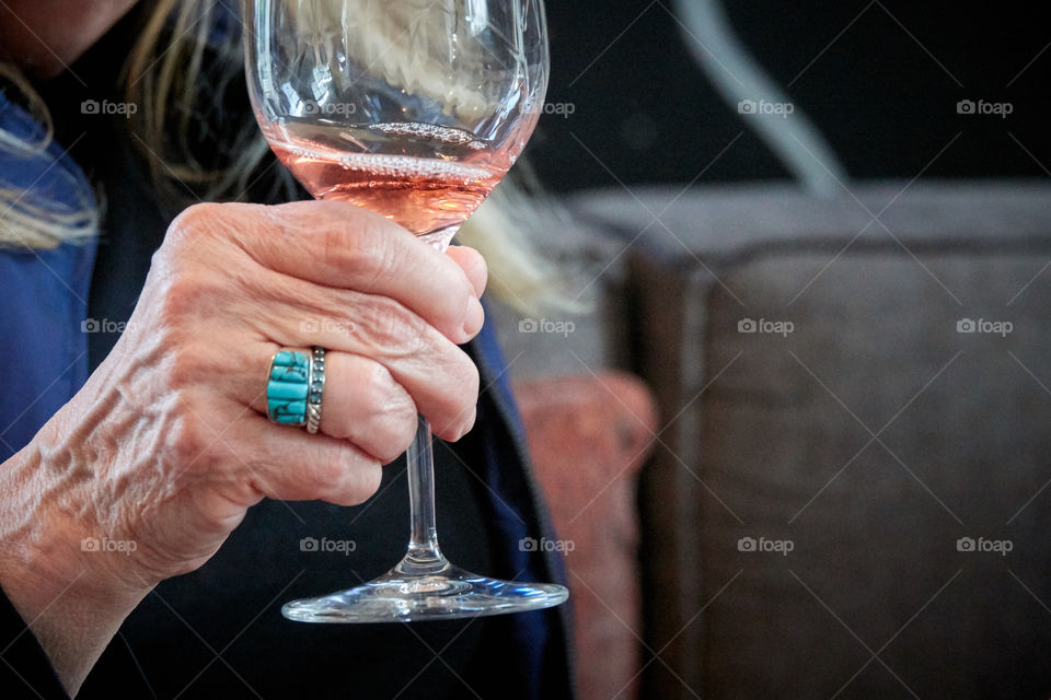 An wealthy old woman’s hand, with a bright blue stone ring, holding a wine glass with rosé wine.