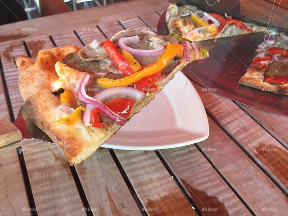 A slice of pizza with colorful toppings being served on a plate at an outdoor restaurant.