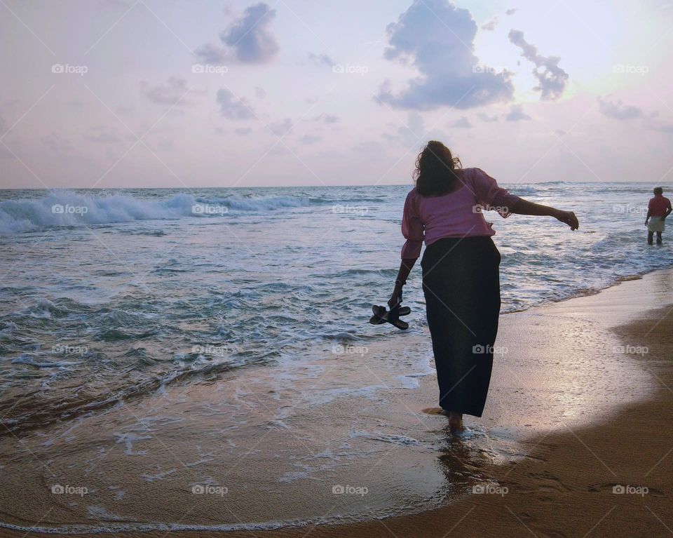 This is a capture of a girl who walking on the golden colour sea beach.She is walking through a shallow wave.There is a crashing wave on the sea.This is a sunsetting scene in the evening and sky is much cloudy and the horizon is clear.