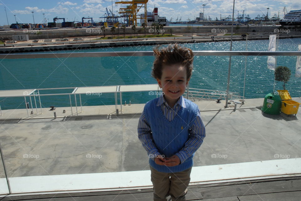 El niño sonríe mientras el viento juega con su pelo con el puerto marítimo de fondo .