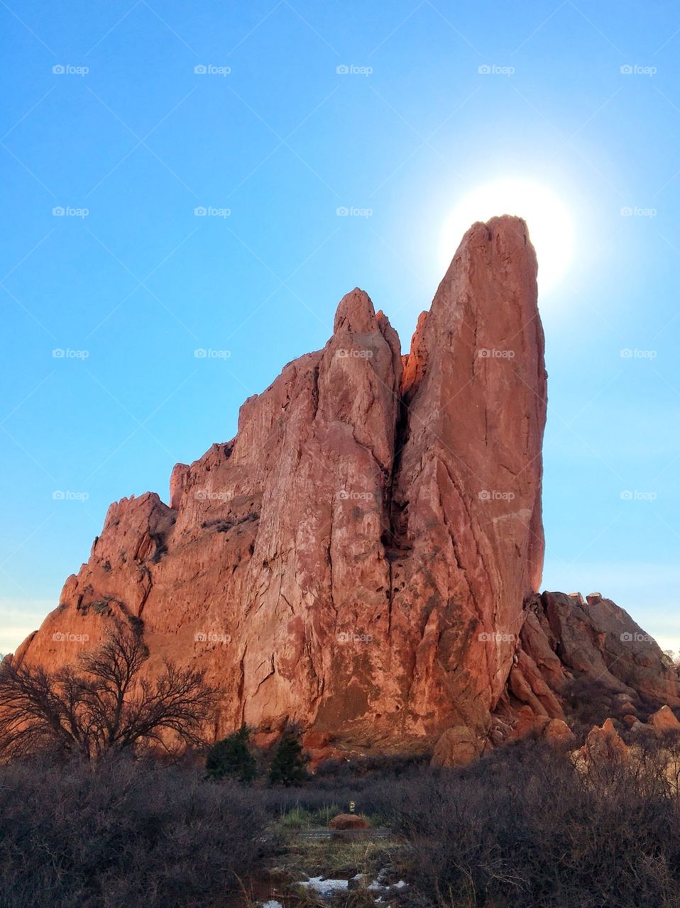 It’s a mid-afternoon day during winter. A red-rock formation is illuminated by the sun that is just barely obscured by one of its apexes. The sky is a true sky blue. There is shrubbery at its base. A bit of snow from the last snowfall lingers.